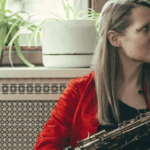 caroline davis - woman sits in front of a dresser covered in pot plants with her saxophone
