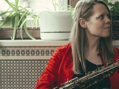 caroline davis - woman sits in front of a dresser covered in pot plants with her saxophone