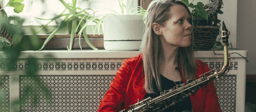 caroline davis - woman sits in front of a dresser covered in pot plants with her saxophone