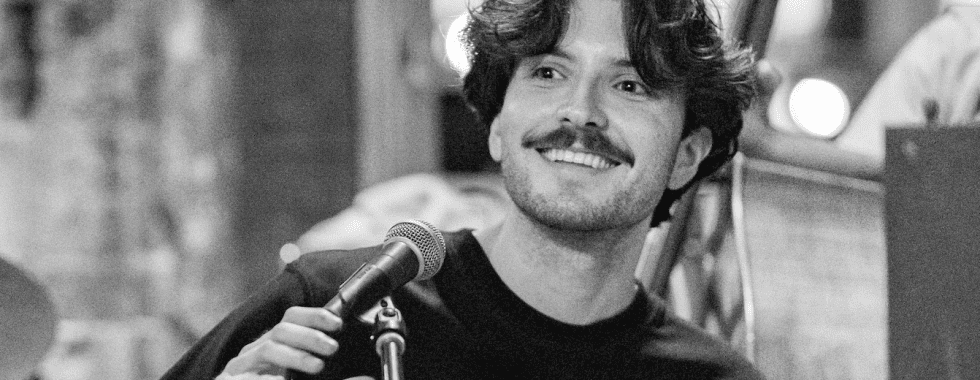 man smiles while holding a microphone and sitting at piano in black and white photograph