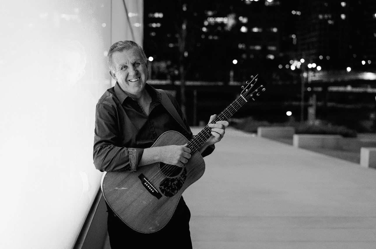 Jamie Clark stands next to a building holding his guitar and smiling